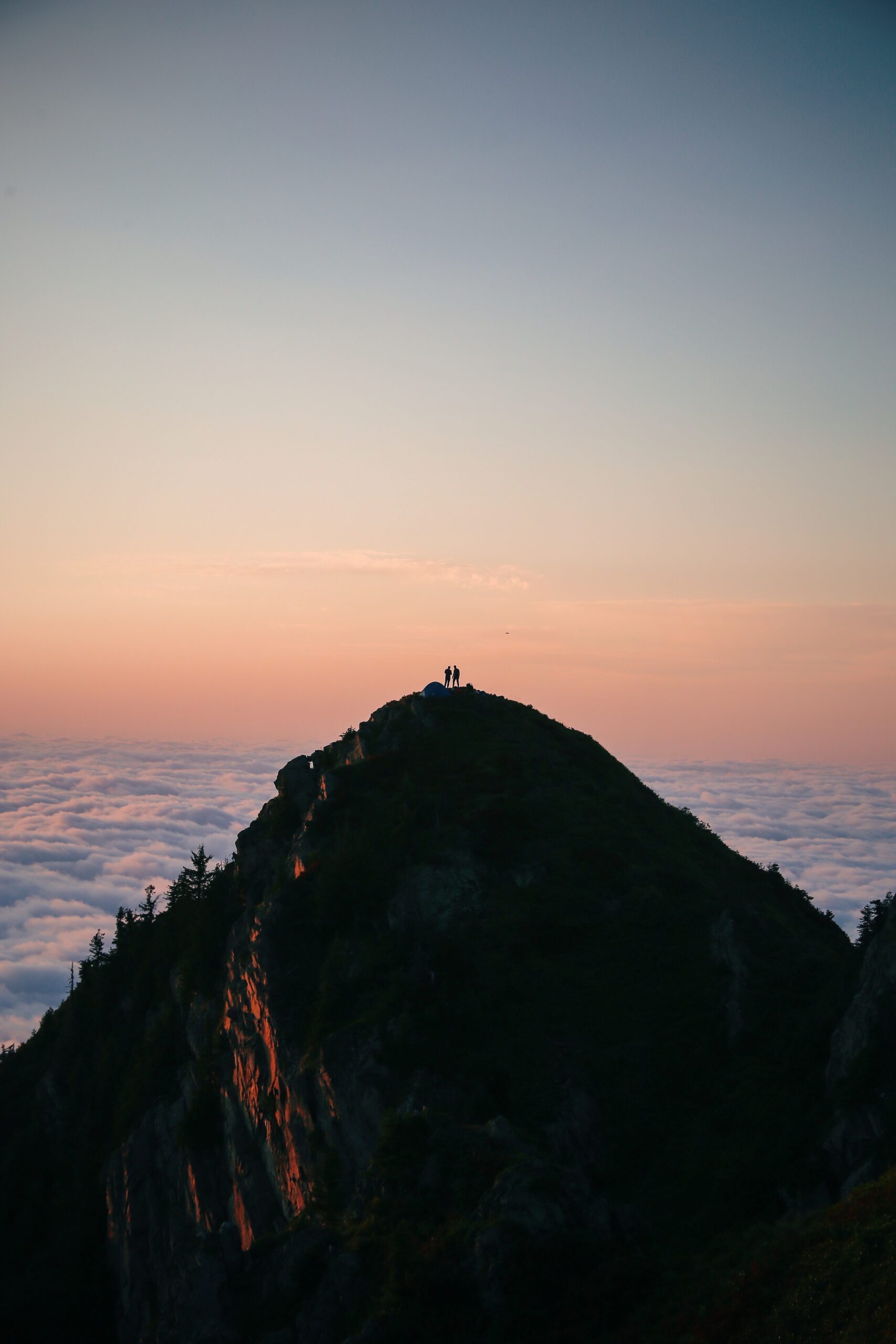 a mountain reaching into the sky with clouds visible below its peak and someone on top of its summit